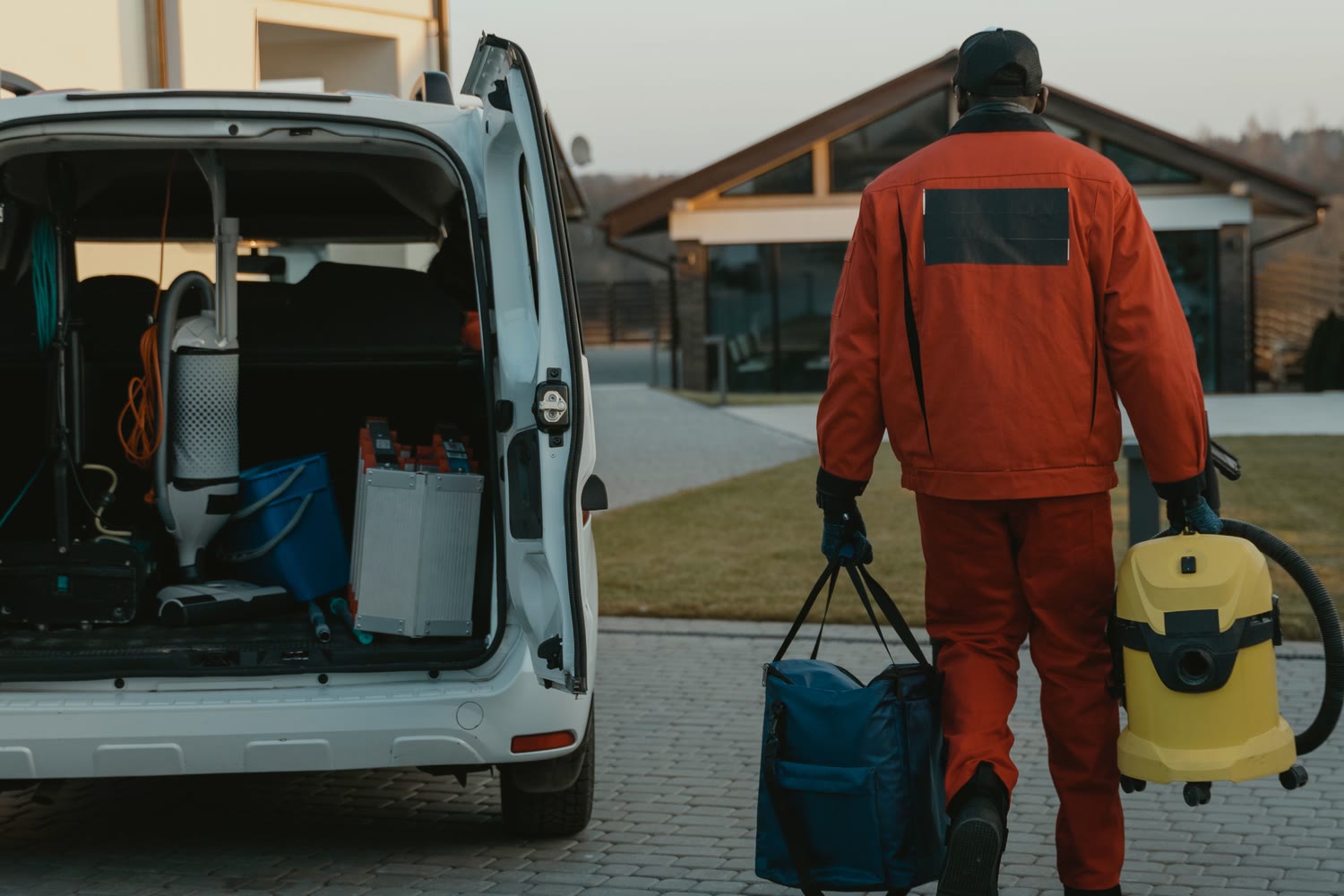 A service contractor carrying a vacuum and equipment bag from an open, fully stocked work van toward a house at golden hour